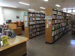 左手前に茶色のカウンター、左奥には長いソファーが設置されており、右奥にはたくさんの本が並んでいる6段の本棚が2列並んでいる長和町図書館の内観写真