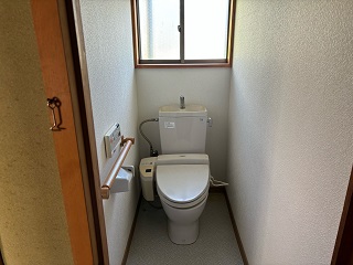 正面奥上部にアルミサッシ、左側にトイレットペーパーホルダー、手摺りが設置された洋式トイレの写真