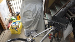 自転車、黄色いカゴ、じょうろ、ビニール傘、脚立などたくさんの物が置かれている玄関の写真