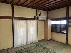 奥には白いカーテンが閉められた窓があり、右奥には茶色の棚が置かれ、畳の上には緑の柄のカーペットが敷かれた和室の内観写真