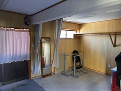 入口には白いカーテンがかけられており、コンクリートの床に木のタイルの壁の店舗で壁には全身鏡が立てかけられ、ハンガーラックが2つ置かれている家の隣に隣接している店舗内装の写真