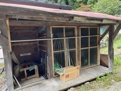 砂利の上の敷地に赤い屋根で木で作られた小屋に収納スペースが3つあり、椅子や農機具、ネギの入った段ボールが置かれている物置の写真