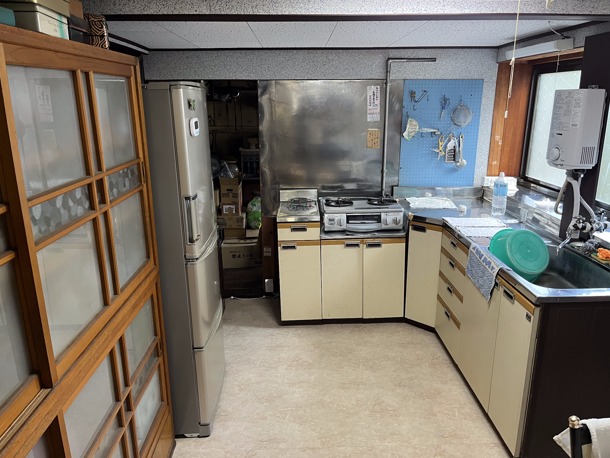 左側に食器棚、冷蔵庫があり、正面奥にガスコンロ、右側に流し台、湯沸かし器、サッシ窓枠がある台所の写真