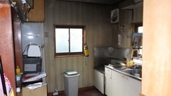 クリーム色の縦ストライプ柄の壁と中央に正方形の窓があり、右にステンレスのキッチン、左に棚や白いトースターが置いてある写真