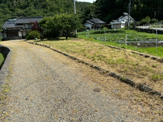 左側に取付け道路、正面奥に樹木、母屋、右側に畑がある地続き農地の写真