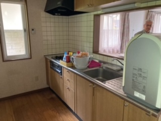 左側奥にサッシ、中央にガスコンロ、調理台、シンクがあるシステムキッチン、右側奥にカーテンが開いたサッシがある、キッチンの写真