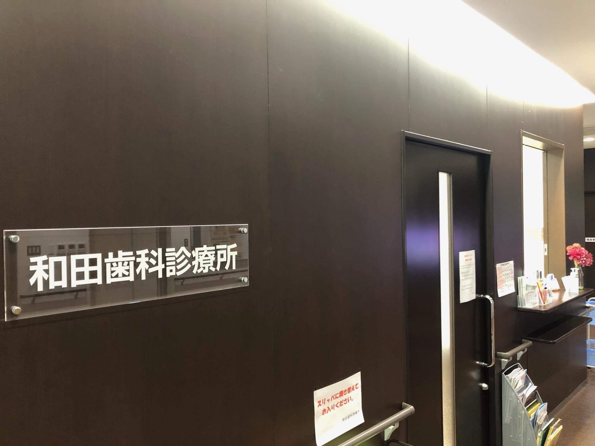 木目調の茶色の壁に和田歯科診療所と書かれたアクリルプレートが設置されており、ドアと受付窓口がある和田歯科診療所の待合室の写真